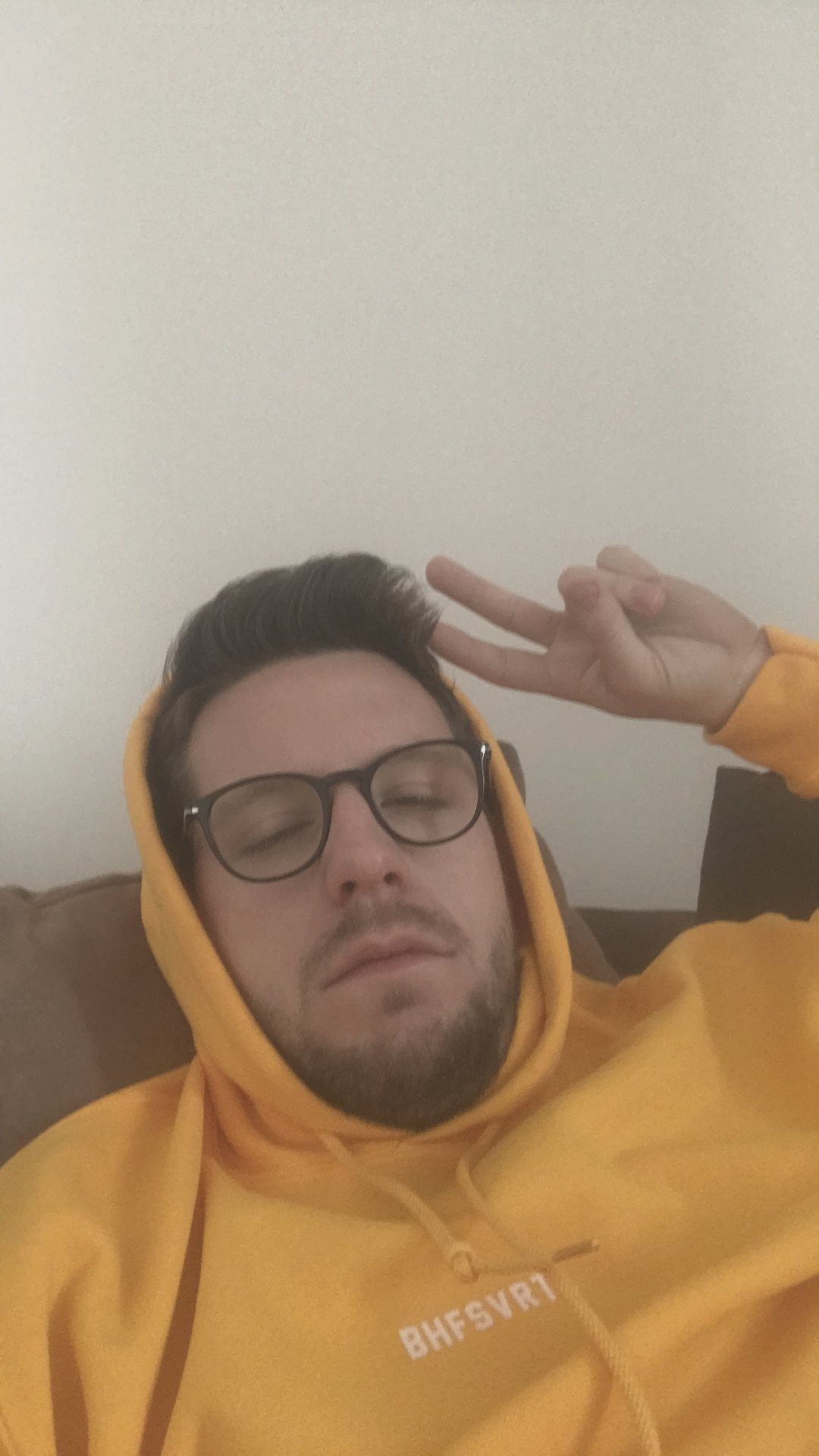 Alex im gelben Hoodie auf dem Sofa, Augen geschlossen, mit Peace-Zeichen